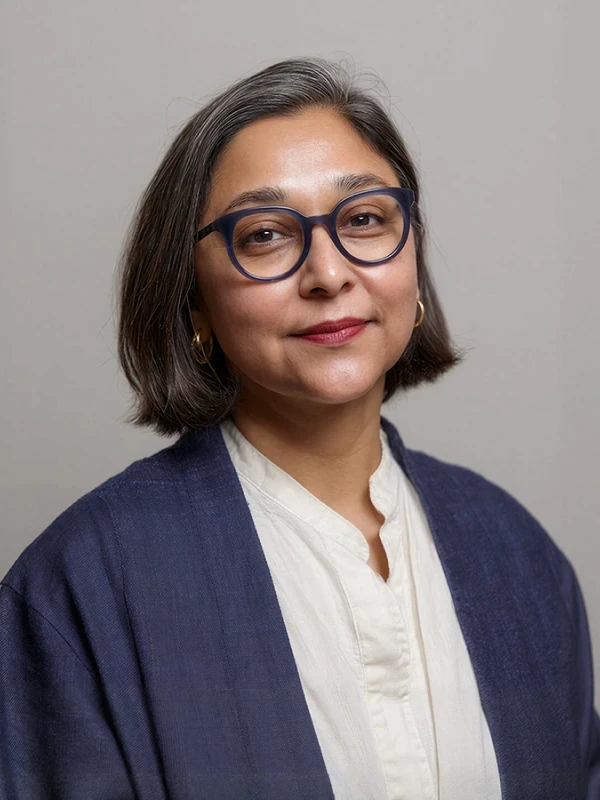 A woman with glasses and dark hair styled in a bob smiles softly, wearing a dark blazer over a light top.
