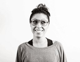 A black and white portrait of a woman facing forward and smiling. She wears round-framed glasses and a dark headband. Her hair is pulled back, and she wears a dark top with a high neckline. The background is plain white.