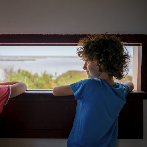 Zwei Kinder schauen aus einem Holzfenster auf eine Landschaft mit Wasser und Grün. Ein Kind trägt ein blaues Hemd, das andere ein rotes.