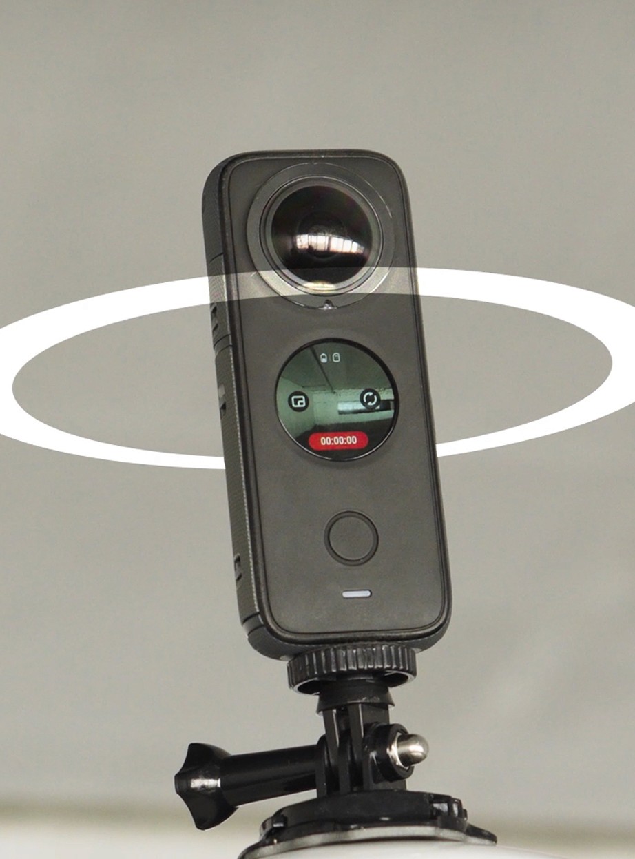Close-up de uma câmera 360° preta montada em um suporte. A câmera possui uma lente circular proeminente e uma pequena tela digital exibindo o status da gravação. Um anel gráfico branco ao redor da câmera simboliza a captura em todos os ângulos simultaneamente.