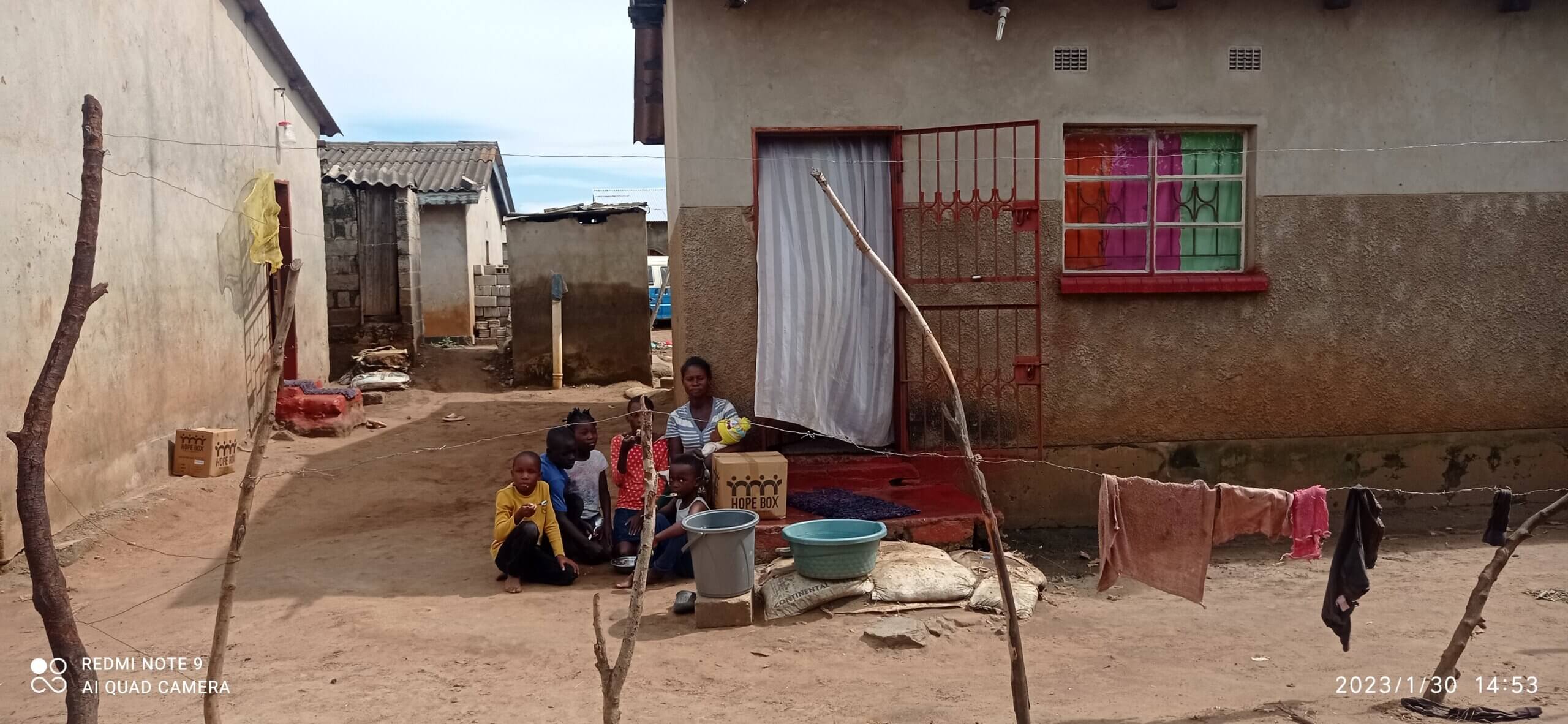 A family in Zambia sits outside their home with Hope Boxes nearby, surrounded by water basins and a clothesline, reflecting the everyday impact of food aid in their community.