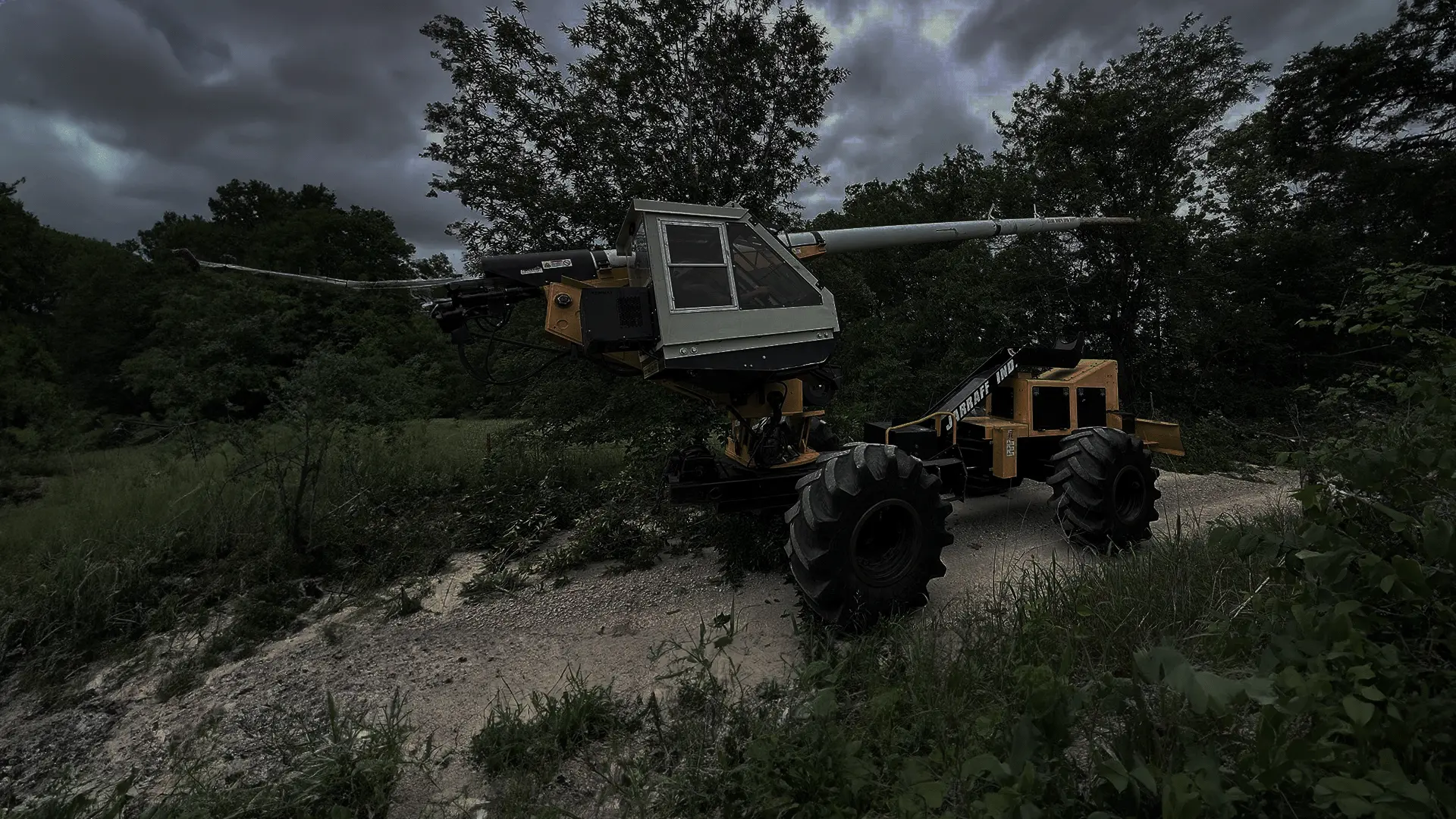 A yellow, tracked vehicle with a large, articulated arm and a bucket at the end. The vehicle is parked on a dirt road in a grassy field with trees in the background. The sky is cloudy.