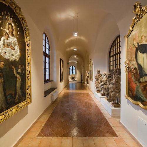 A museum corridor with large religious paintings, statues, and arched windows under a vaulted ceiling.