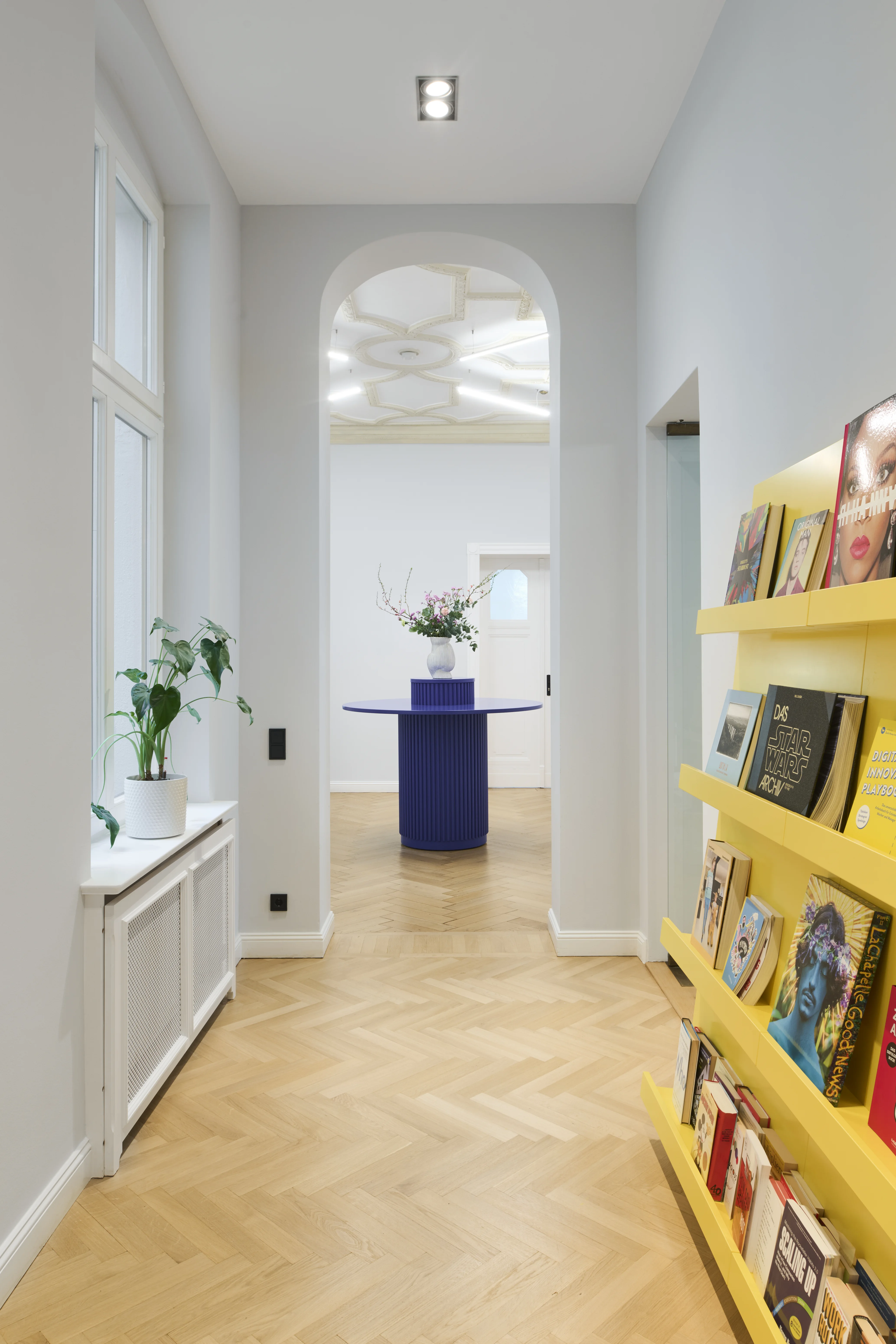 Hallway at INTERMATE BERLIN with herringbone parquet, a yellow wall-mounted magazine rack full of books, plants on a white sideboard and a view through an arched opening to the blue round table.