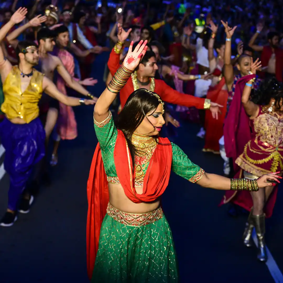 A lively parade with dancers in vibrant traditional attire, featuring a woman in a green and red outfit leading. The scene is festive and colorful.