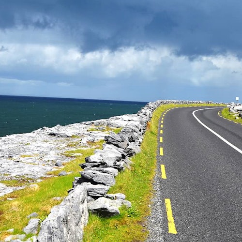 Крайбрежен път с жълти прекъснати линии се извива покрай скалист терен под облачно небе, съседен на синьо море.