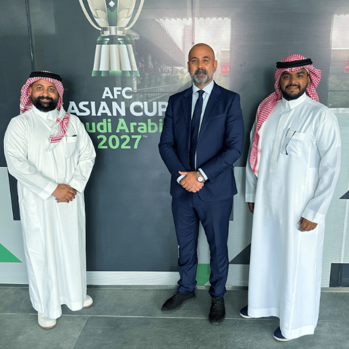 Three men standing before AFC Asian Cup Saudi Arabia 2027 promotional backdrop.