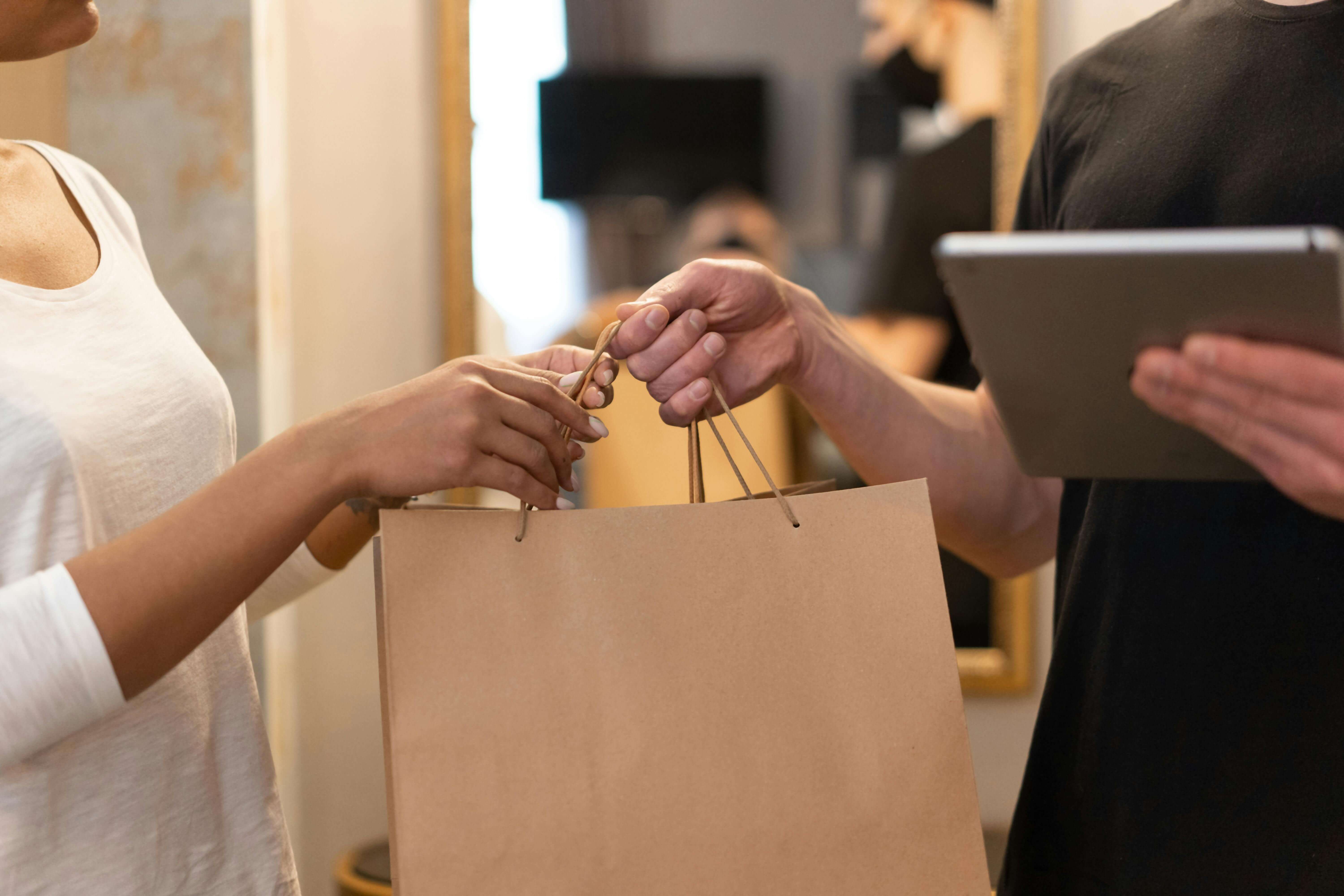 A paper shopping bag exchanging hands. The receiver is using two hands to accept the bag and the giver is behind a electronic tablet.