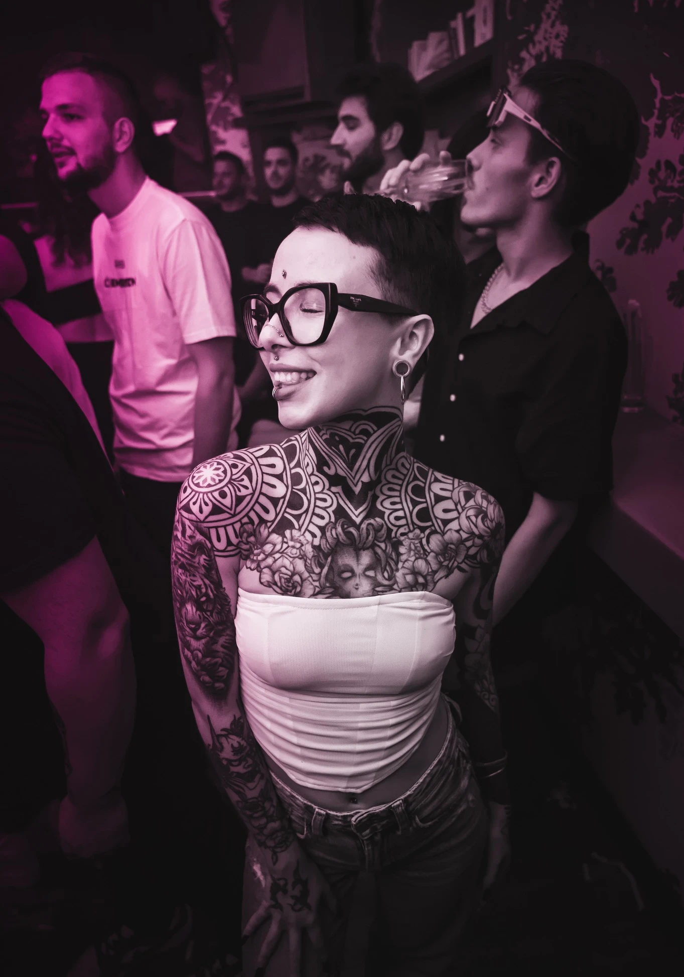 A person with extensive tattoos on their neck, chest, and arms smiles with their eyes closed. They are wearing glasses and a white tube top, standing in a crowded room with pink lighting.