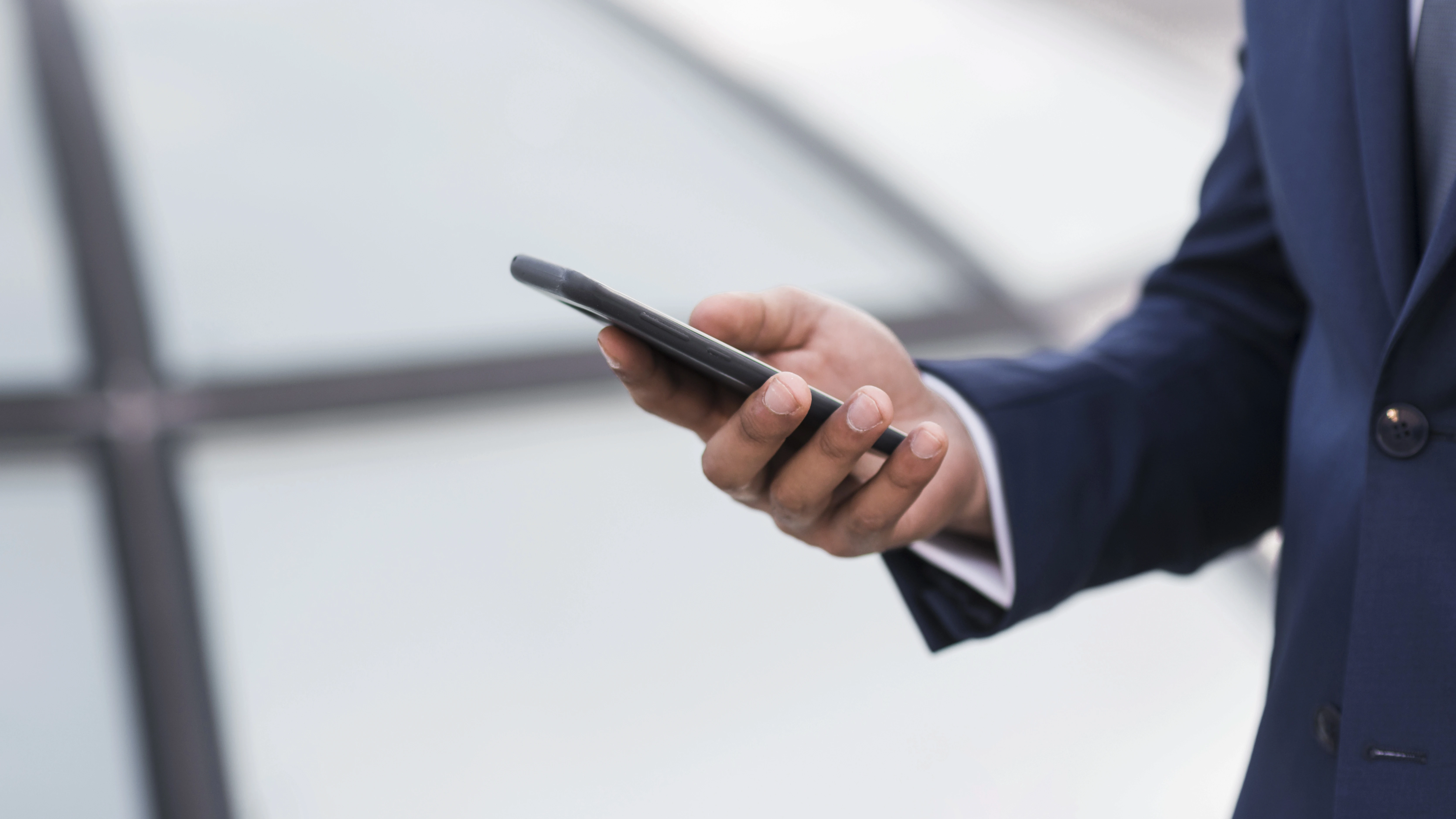 Close-up das mãos de um homem vestindo terno azul segurando e utilizando um smartphone escuro, com um fundo desfocado de arquitetura moderna.