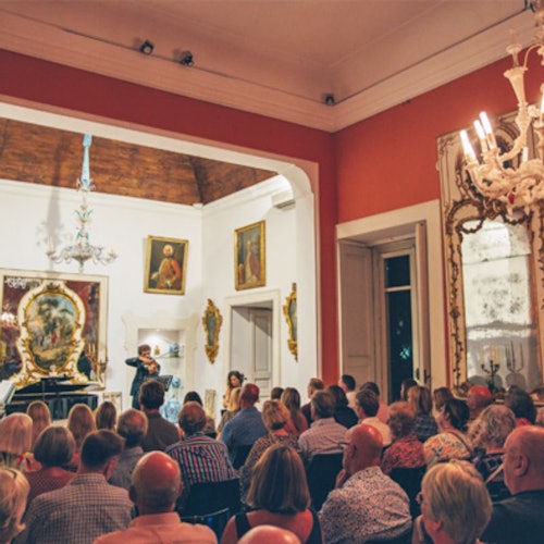 A woman performs on a violin in front of an audience inside an elaborately decorated room with paintings and chandeliers.