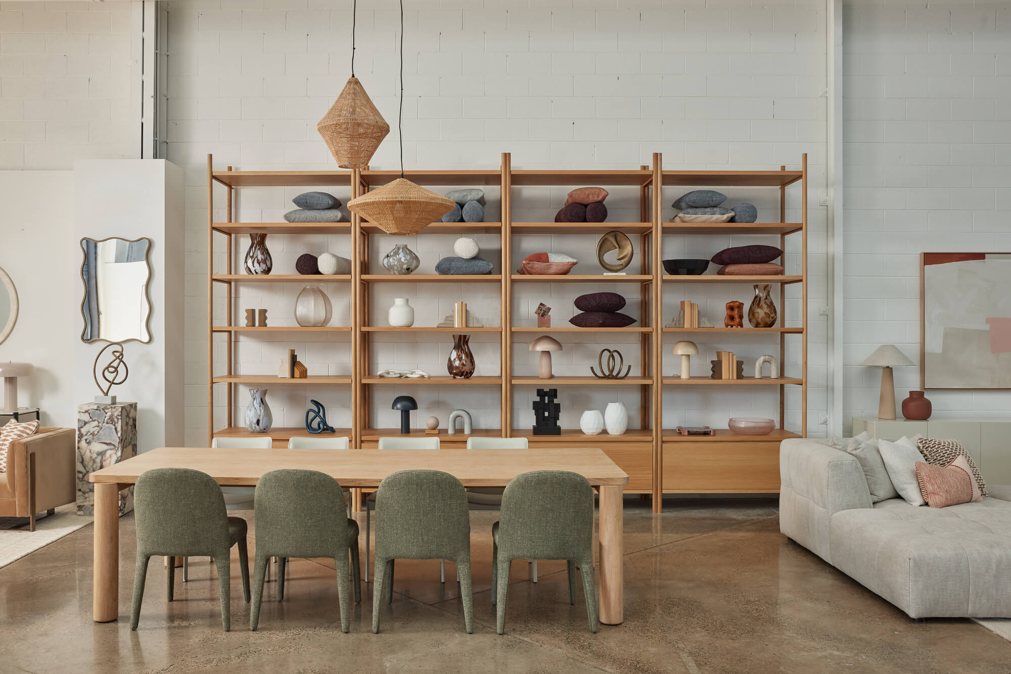 Contemporary wooden dining table, green upholstered chairs, woven pendant lamps, and large oak bookshelf unit in a modern furniture showroom.