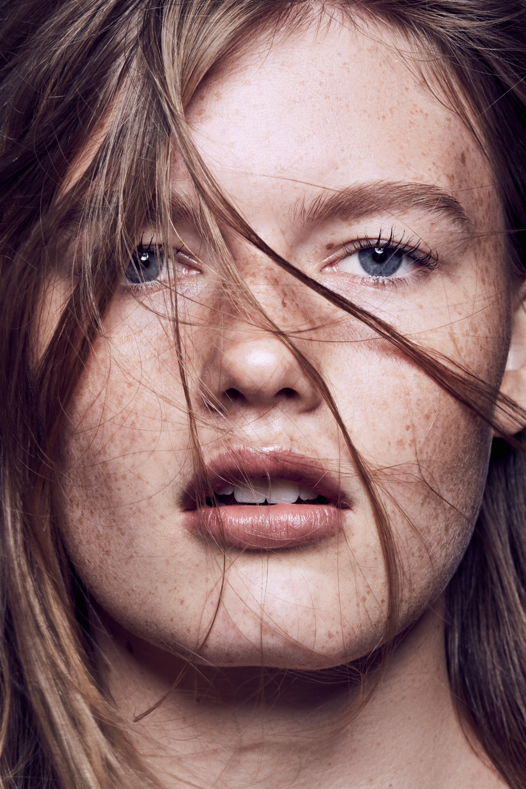 Close-up beauty photography of fair-skinned model with blue eyes and freckles looking straight to camera with her mouth slightly open strands of her mouse-brown hair blown across her face.