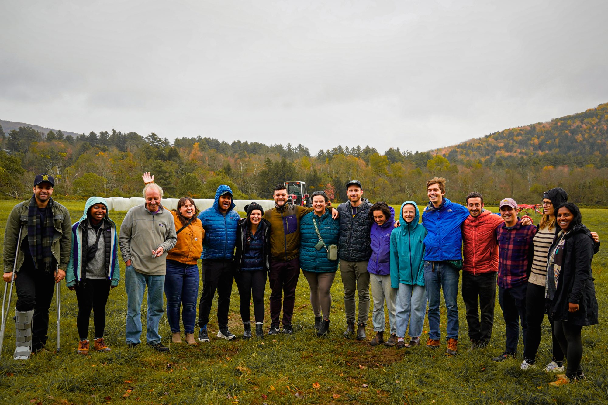 A diverse group of people stands together outdoors, wearing colorful jackets, with mountains in the background.