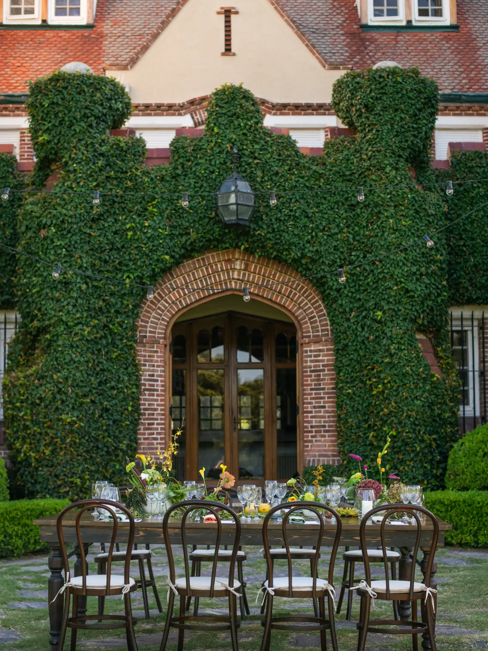 Mesa larga con arreglos florales coloridos frente a fachada con hiedra para evento elegante de inspiración europea.