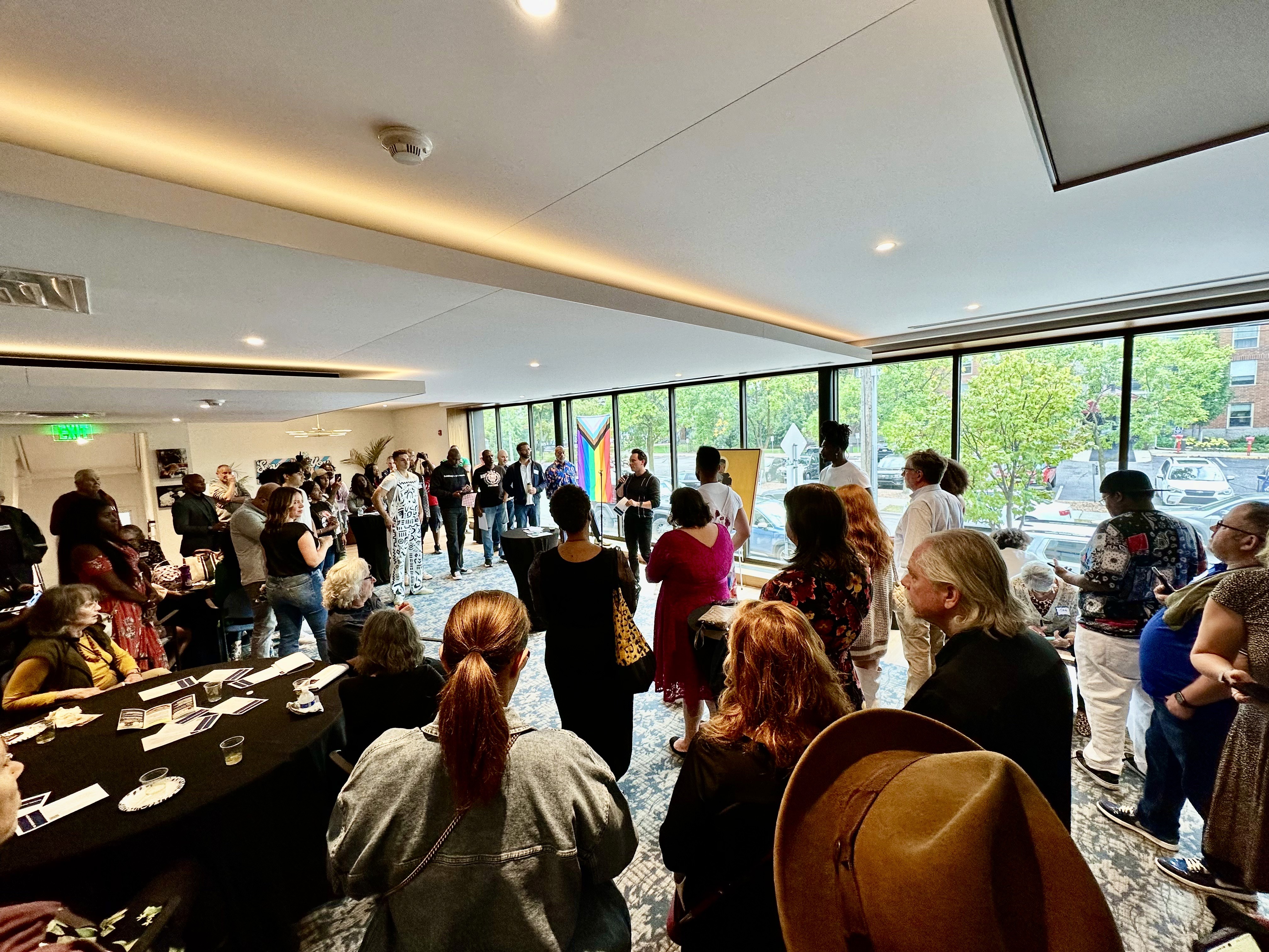 A large group of people gathered in a bright room with floor-to-ceiling windows at Grace Trinity Church, listening to a speaker. A Pride flag is visible in the background.