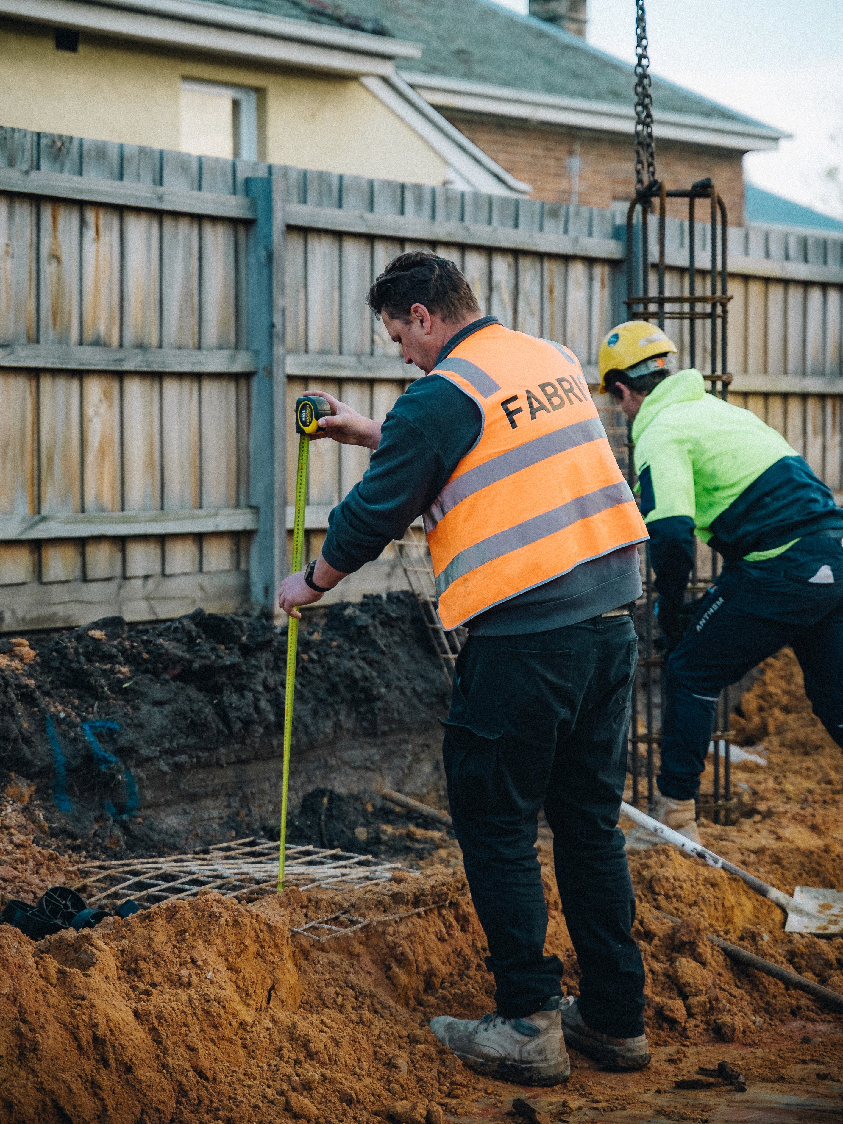 Construction workers measuring foundation depth with tape measure at building site.
