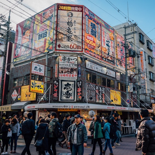 人来人往的城市街景中，人们走过一个建筑物的拐角处，该建筑物上覆盖着许多色彩斑斓的日文招牌。