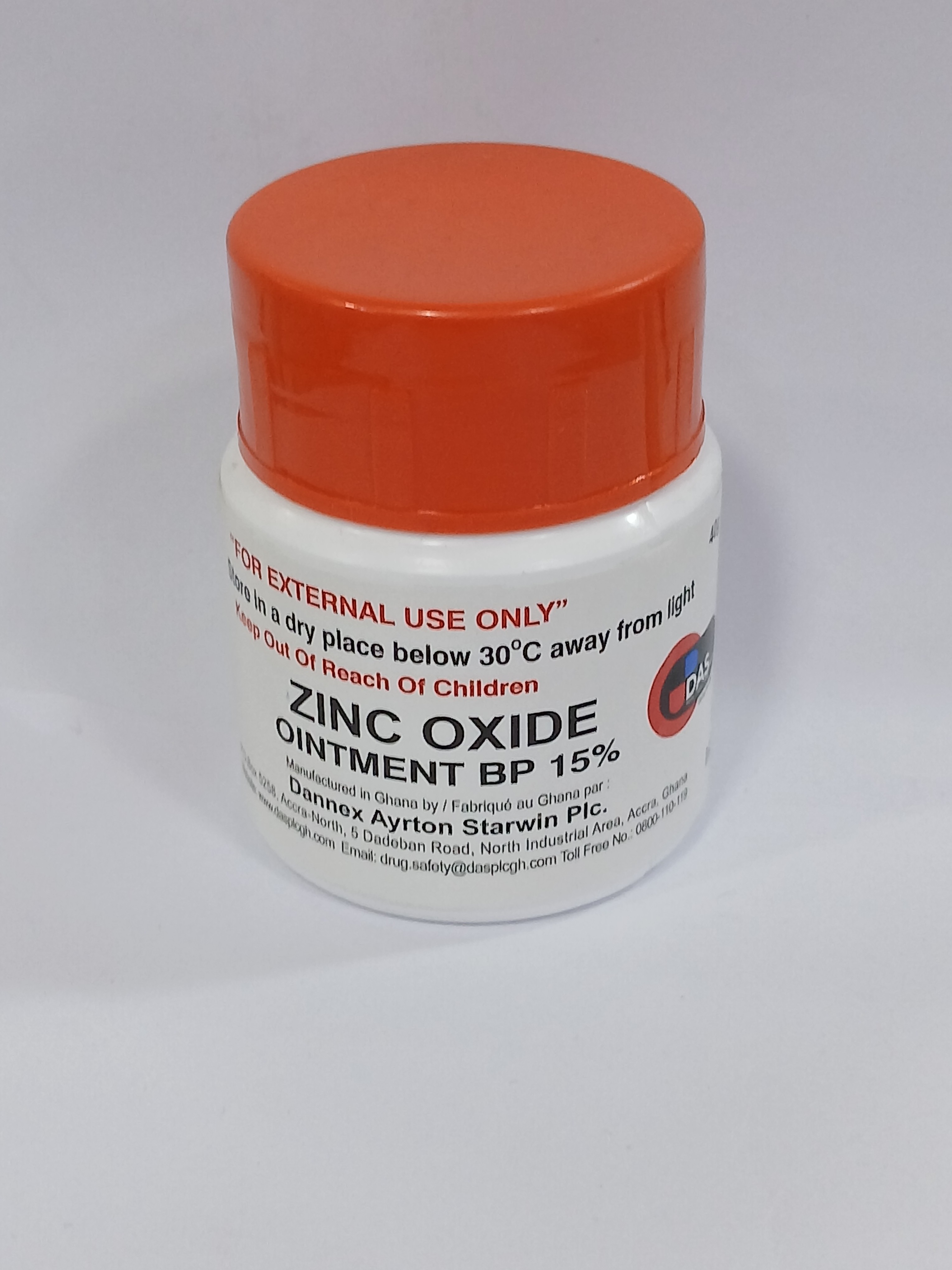 A small white jar with an orange screw-top lid sits against a plain background, featuring printed text in black and red warning labels.