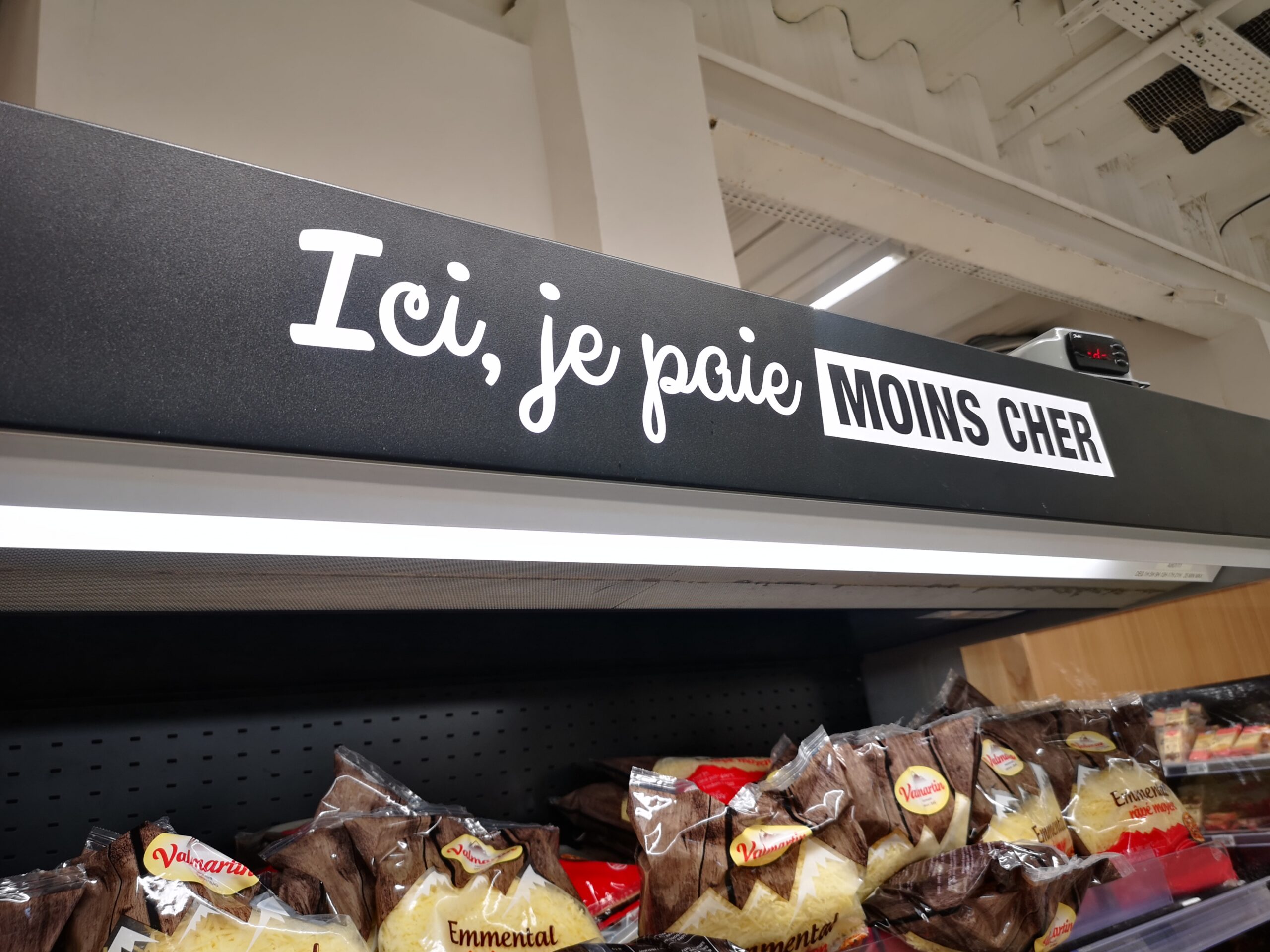 Signalétique de Rayon de supermarché avec une affiche indiquant « Ici, je paie moins cher » en français. En dessous, des sachets de fromage râpé sont exposés.