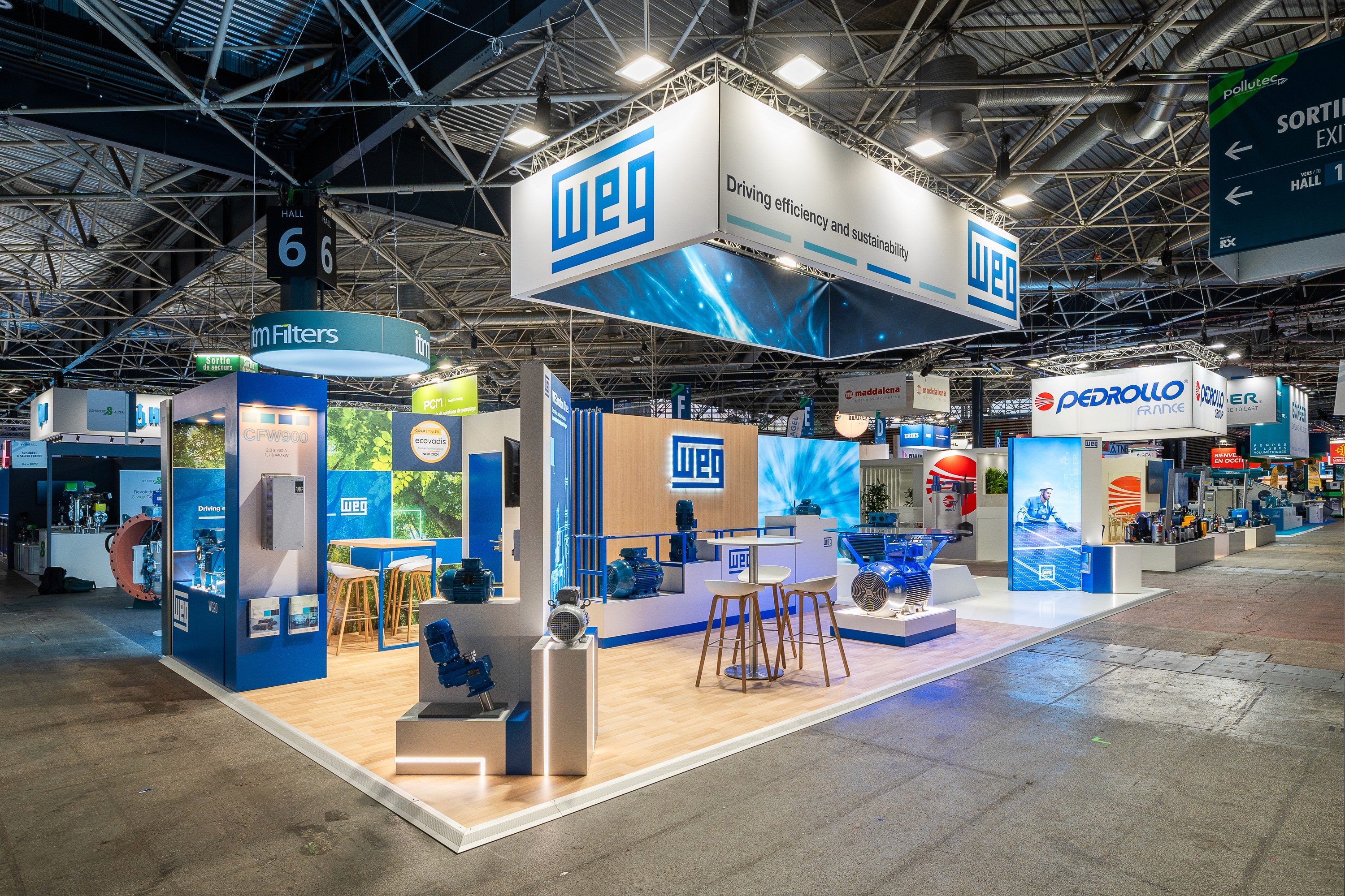 Stand WEG Pollutec 2025 réalisé par Expace – vue frontale du stand avec bandeau lumineux Driving efficiency and sustainability, moteur industriel sur podium vitré rétroéclairé, grands écrans LED et espace lounge bois