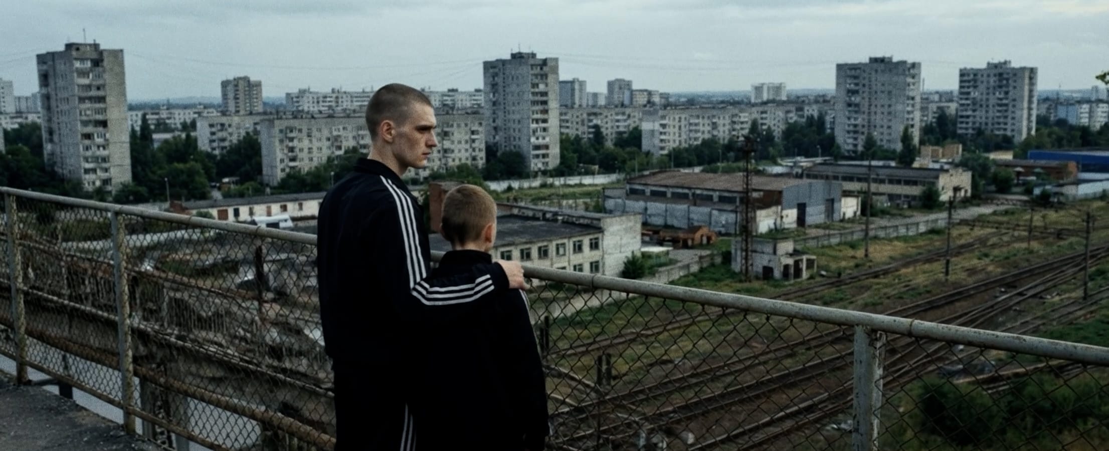 Two figures stand on a bridge overlookin a vast Soviet-era cityscape. Both wear tracksuits; the older puts his hand on the younger's shoulder.