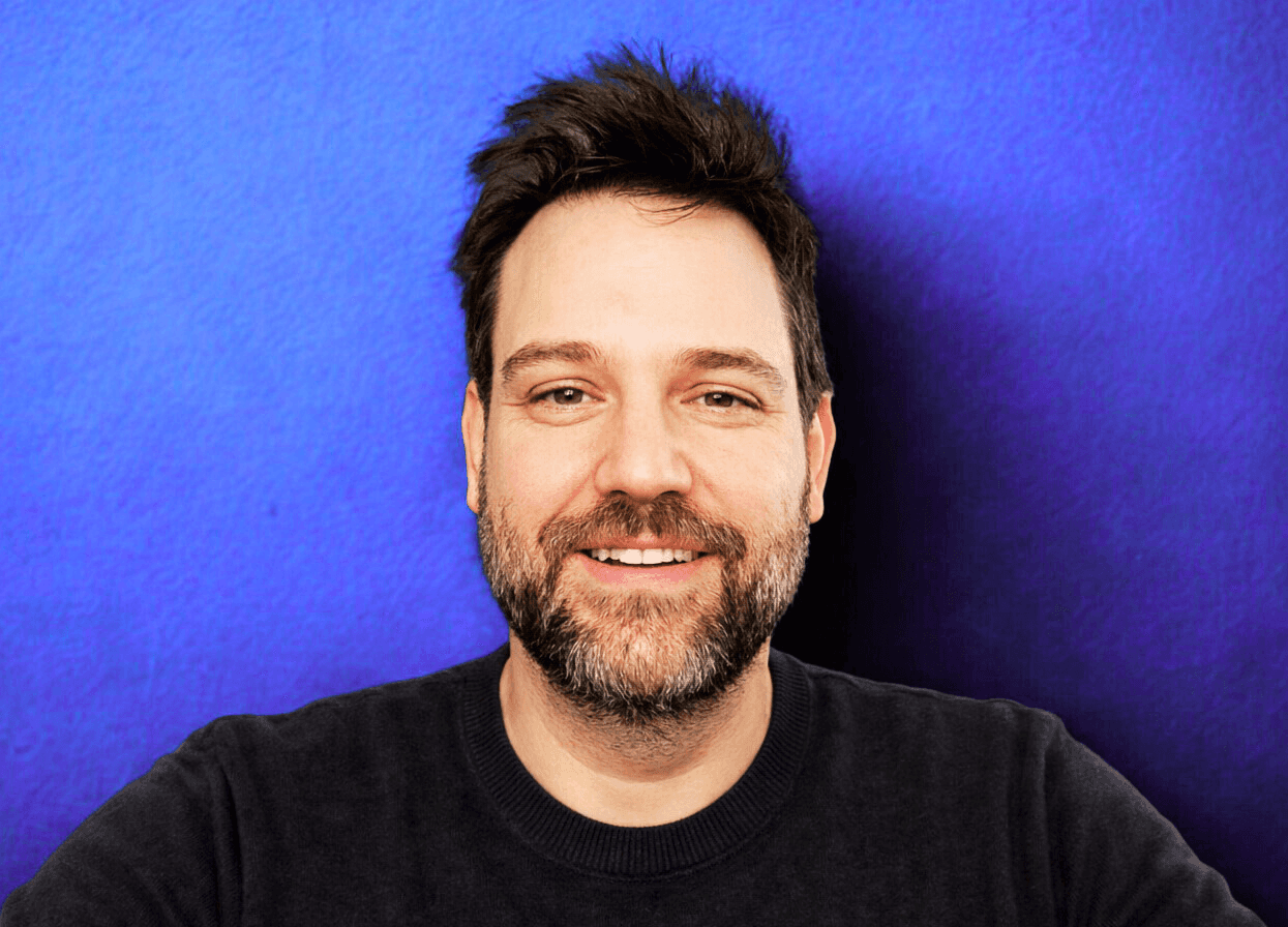 A smiling man with curly hair is sitting at a desk, looking at the camera, with a laptop and a lamp in the background.
