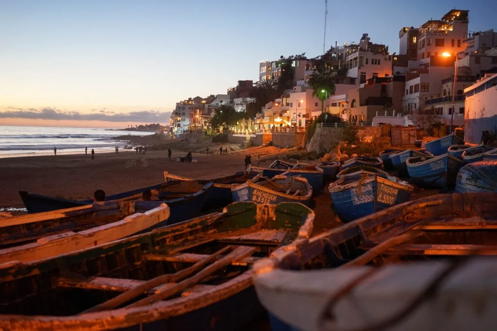 Wide view of Taghazout at dusk, fishing boats resting along the shore below the village