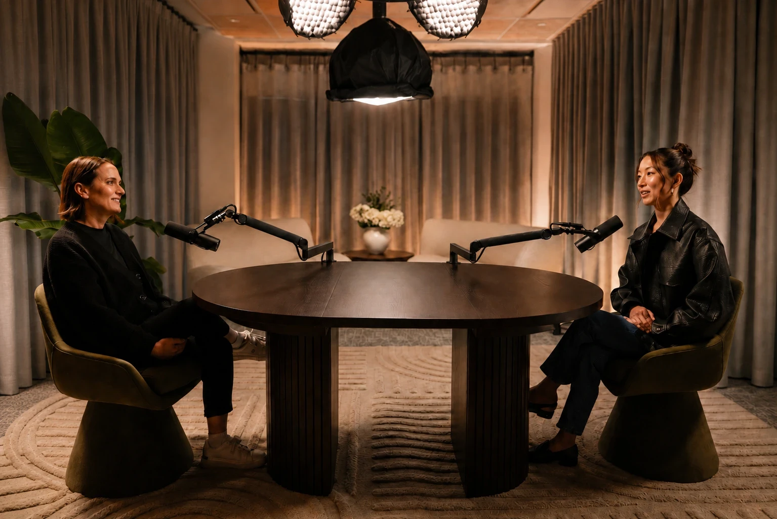 Two people mid-conversation at a round table with microphones in a warmly lit Melbourne studio