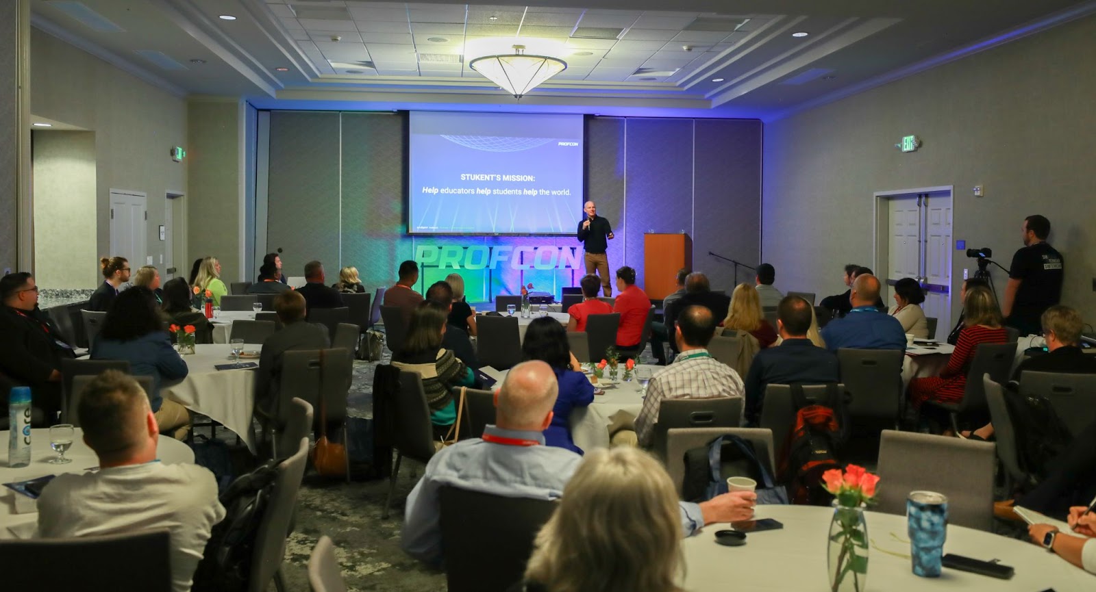 Speaker addresses a full audience at PROFCON conference. Slide behind reads: “STUKENT’S MISSION: help educators help students help the world.” Attendees sit at round tables with flowers, drinks, and notebooks.