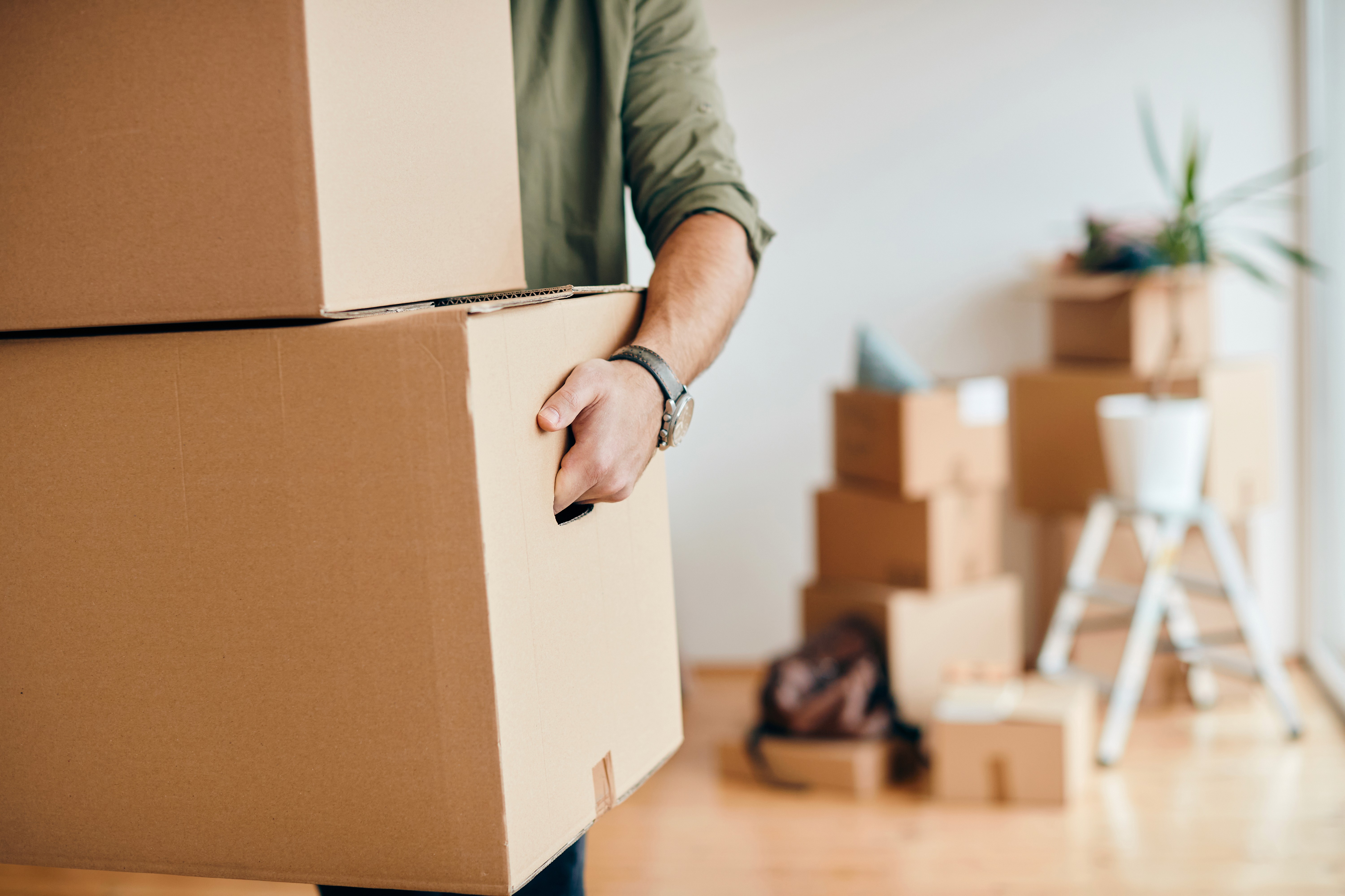 Close-up of a person carrying heavy cardboard moving boxes with more packed boxes blurred in the background.