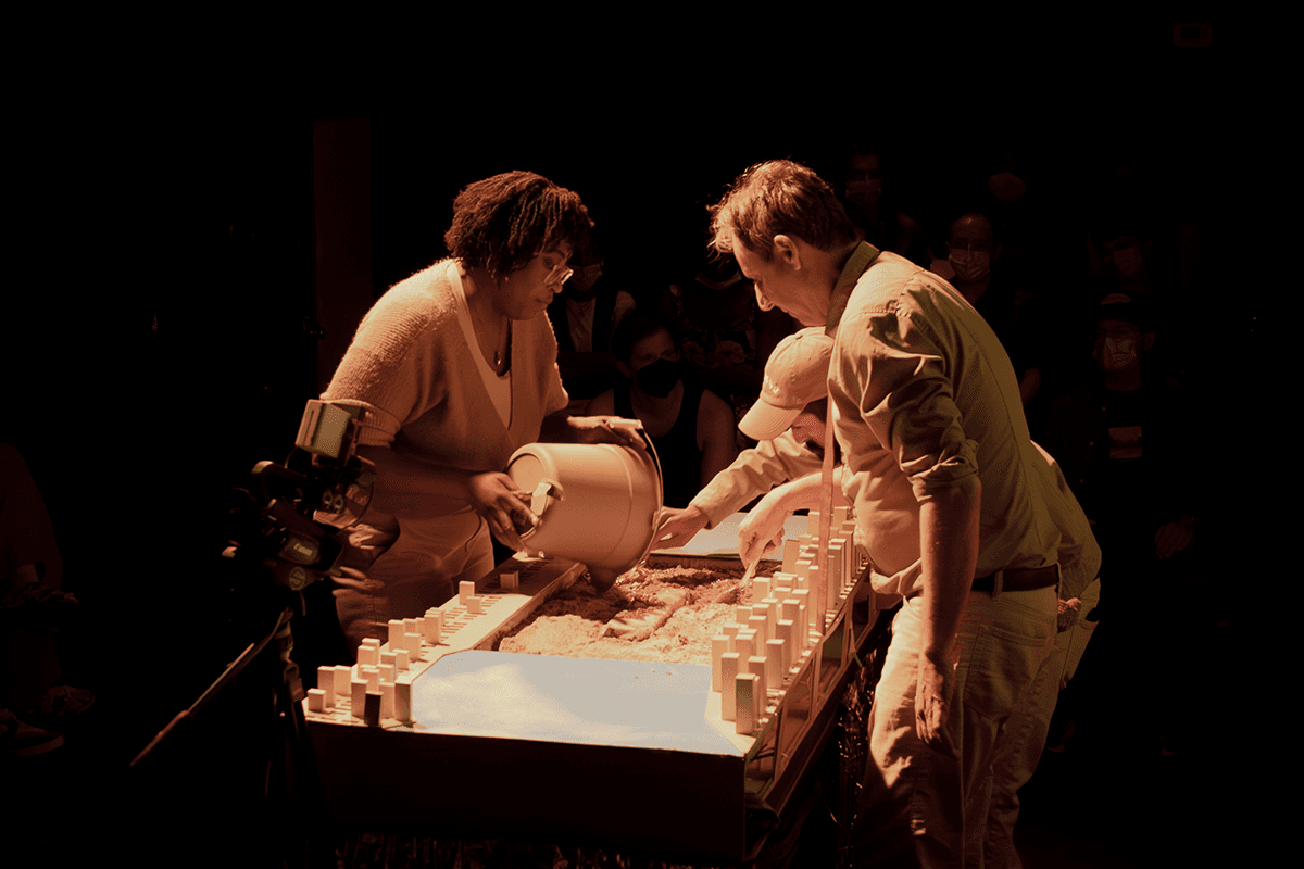 Three performers lean over a miniaturized East River with a bucket and some spoons.