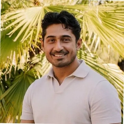 Portrait of a young man in a brown coat and beige turtleneck, with light brown hair and a calm expression against a gray background.