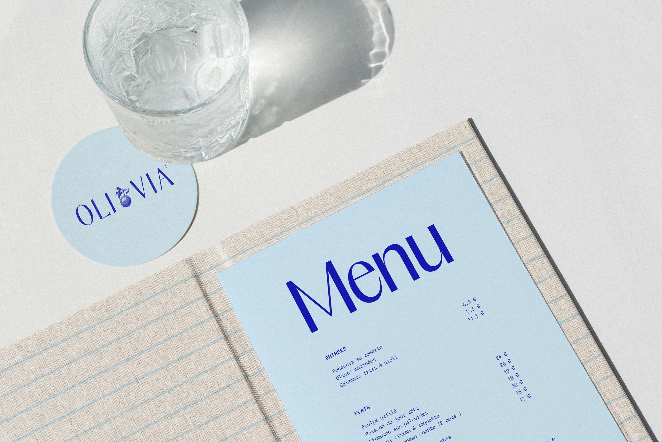 Menu du restaurant OLI•VIA bleu vif sur table avec sous-verre illustré, papeterie haut de gamme pour restaurant méditerranéen contemporain.