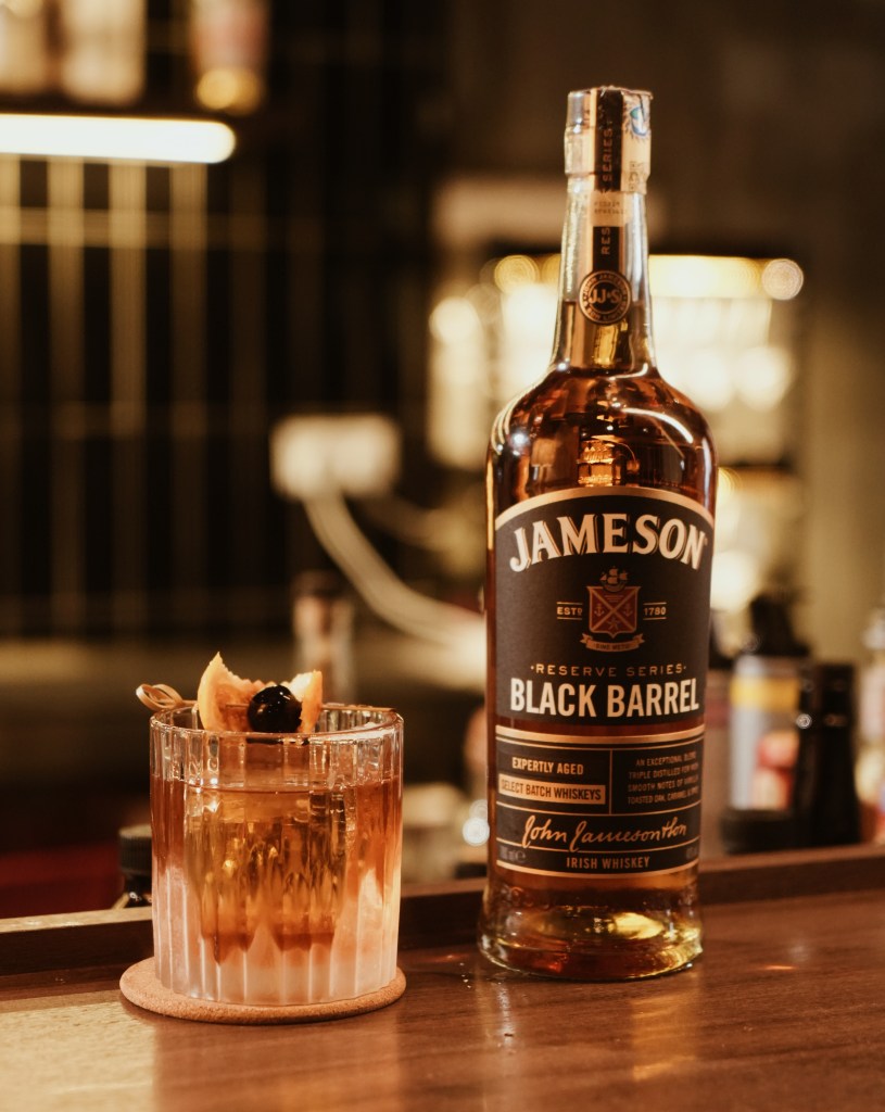 A bottle of Jameson Black Barrel whiskey next to a glass filled with an aromatic cocktail, garnished with orange and a cherry, presented on a wooden surface.