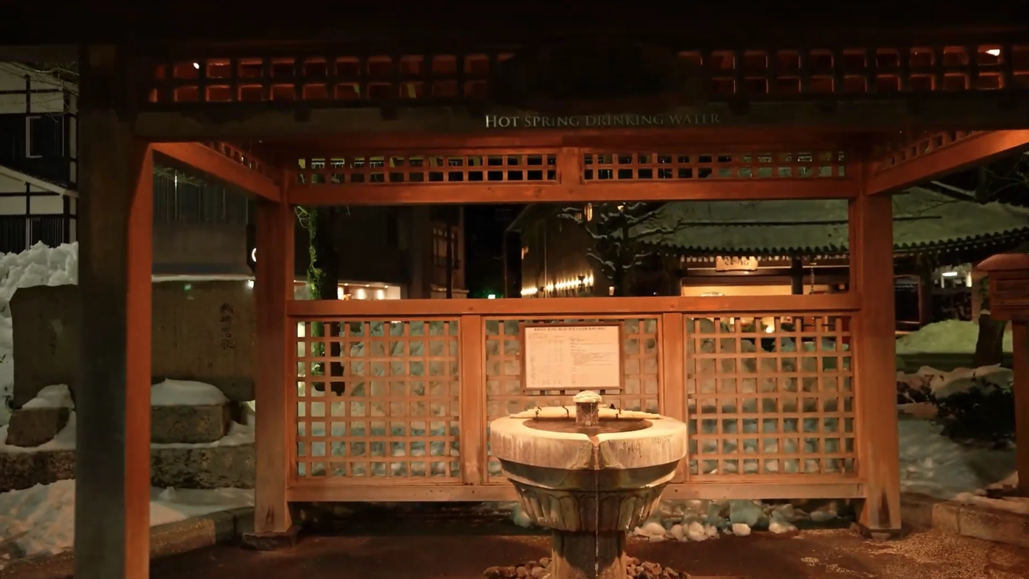 The hot spring drinking pavilion lit at night with snow banked around its stone basin.