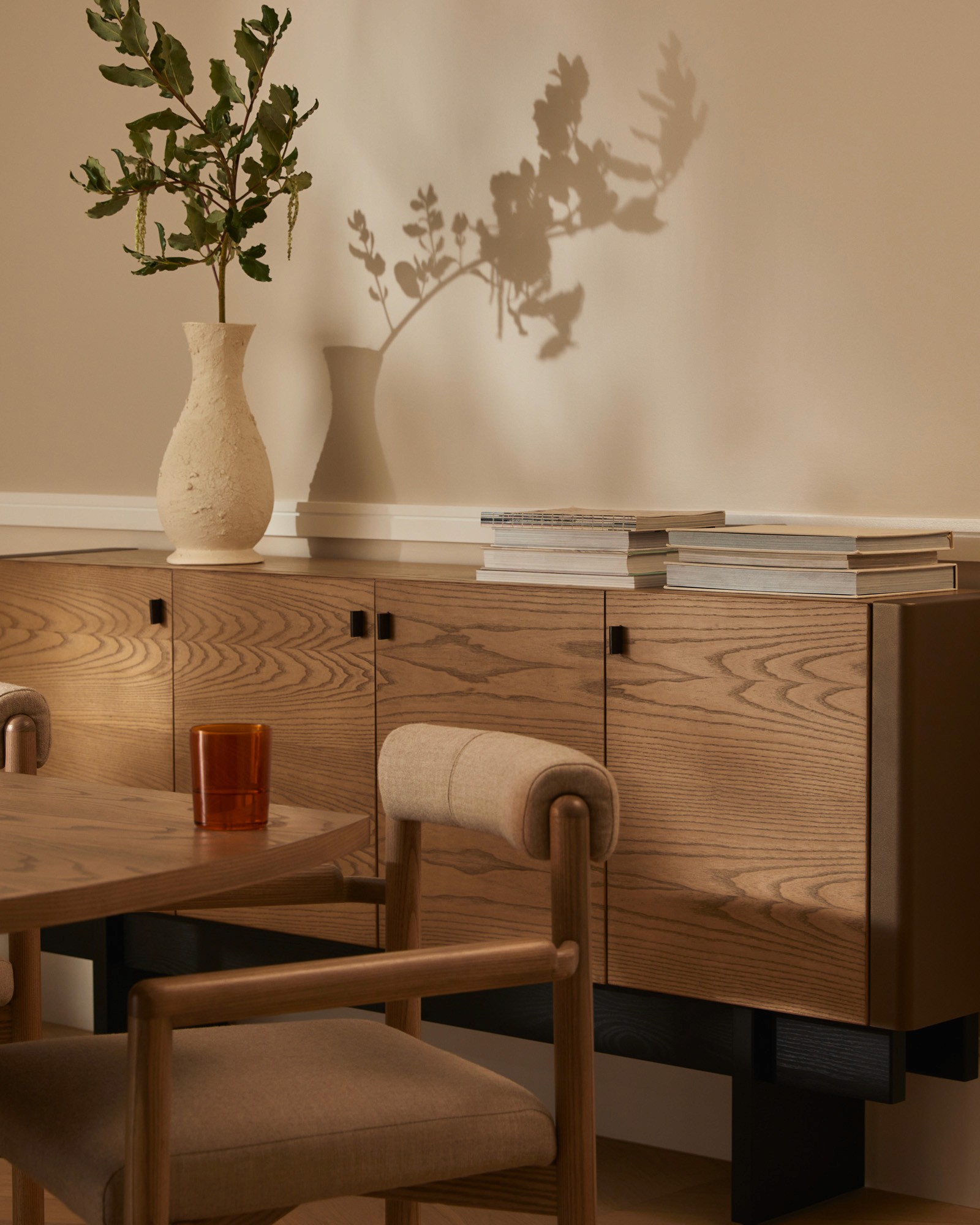 Wooden sideboard with a ceramic vase and plant, stacked magazines and books, and a dining chair in front