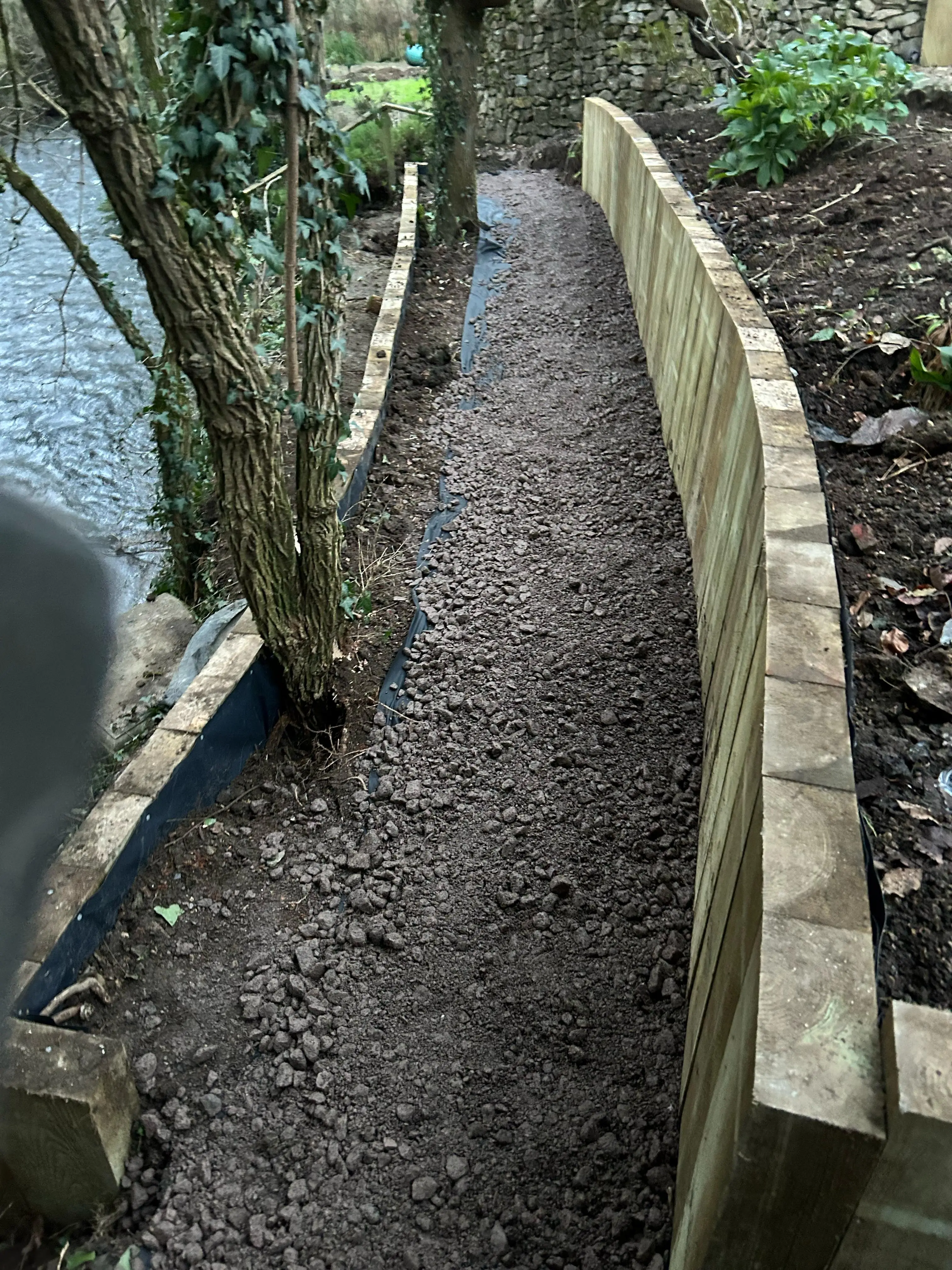 A narrow, winding path alongside a stone wall, surrounded by greenery and soil, next to a body of water.