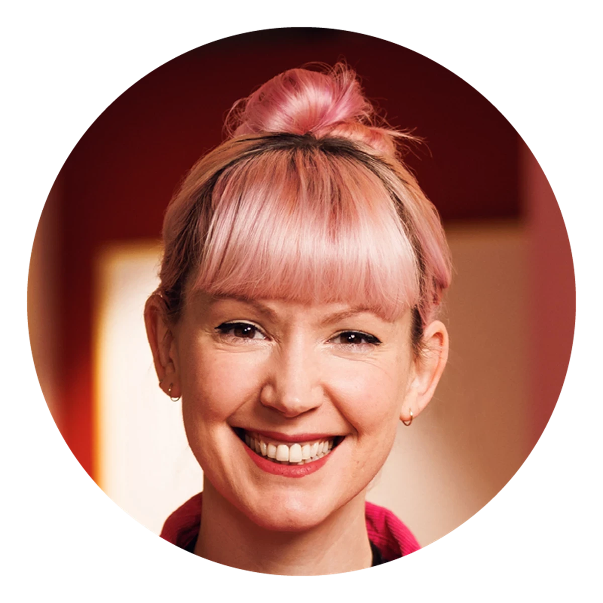 A circular portrait shows a person with pink hair styled in a top knot. Warm indoor lighting and a red background create a bold, modern feel.