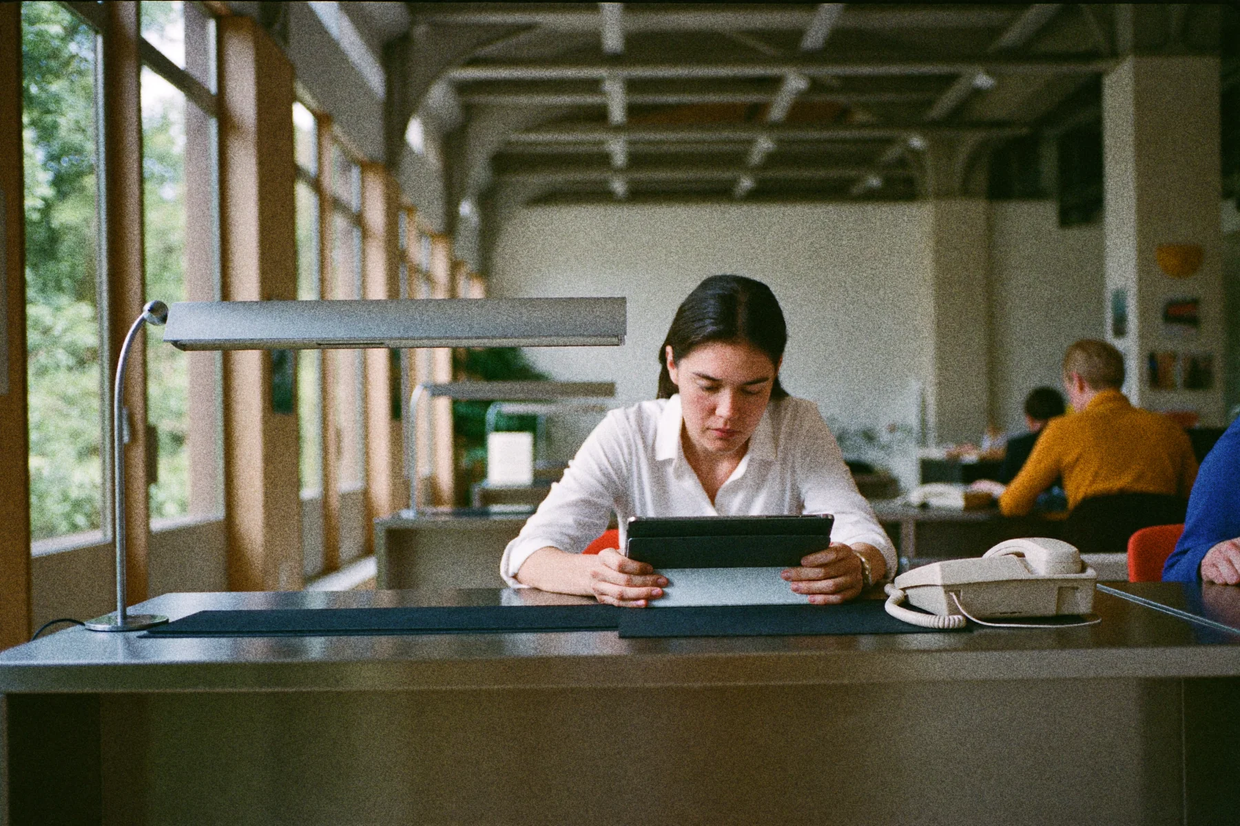 Een receptioniste bekijkt een tablet op een kantoor aan de balie, met een vaste telefoon naast haar op het bureau.