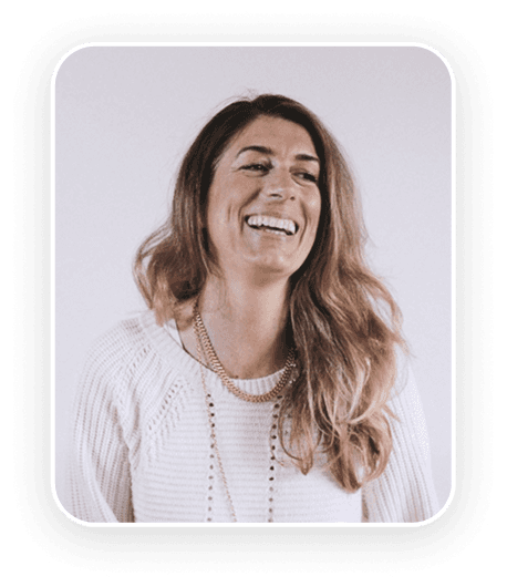 A smiling individual with long hair wearing a light-colored top against a neutral background.