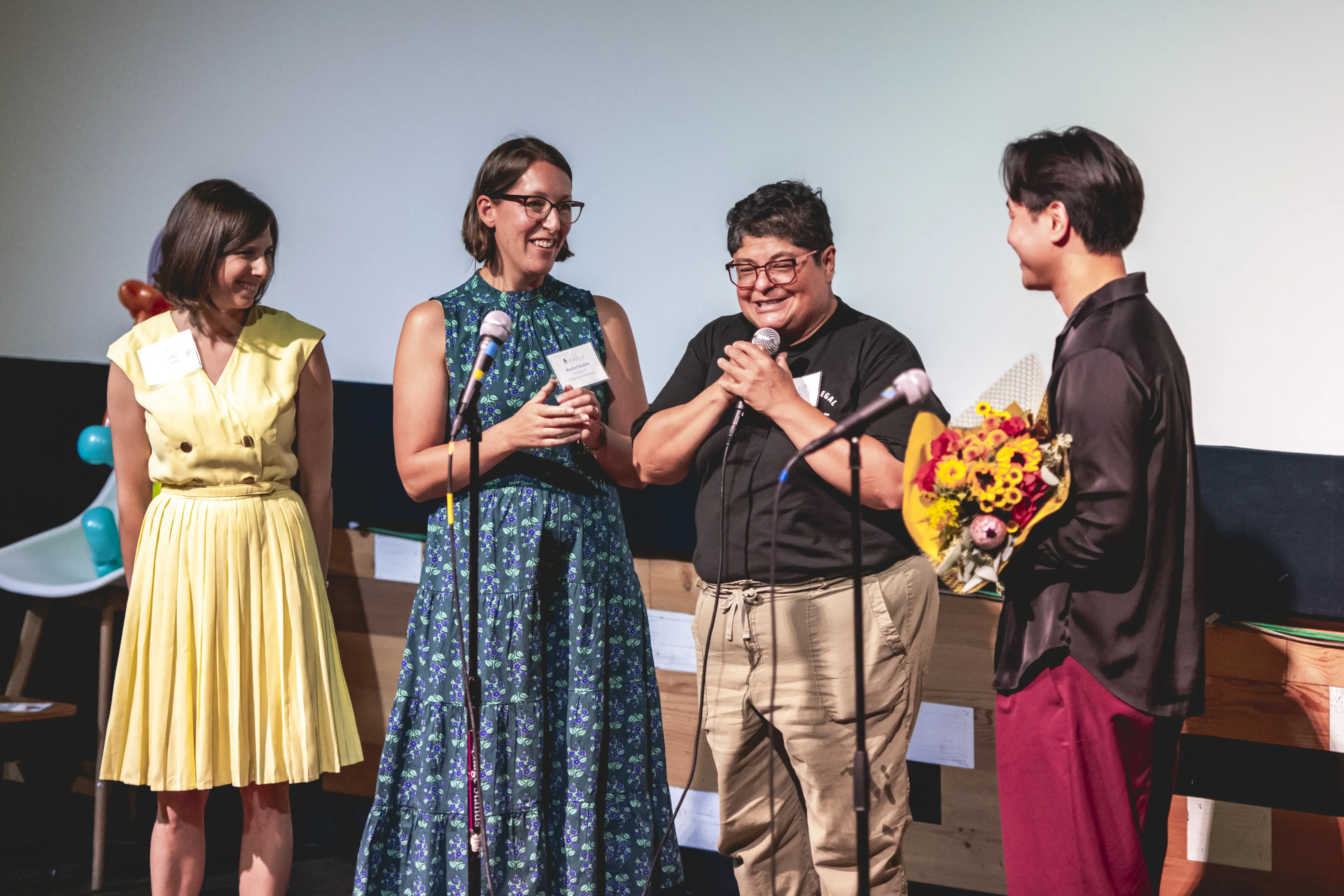 Um grupo de quatro pessoas está em um palco com microfones, sorrindo e rindo, enquanto uma pessoa segura um buquê de flores coloridas, diante de uma tela de projeção branca ao fundo.