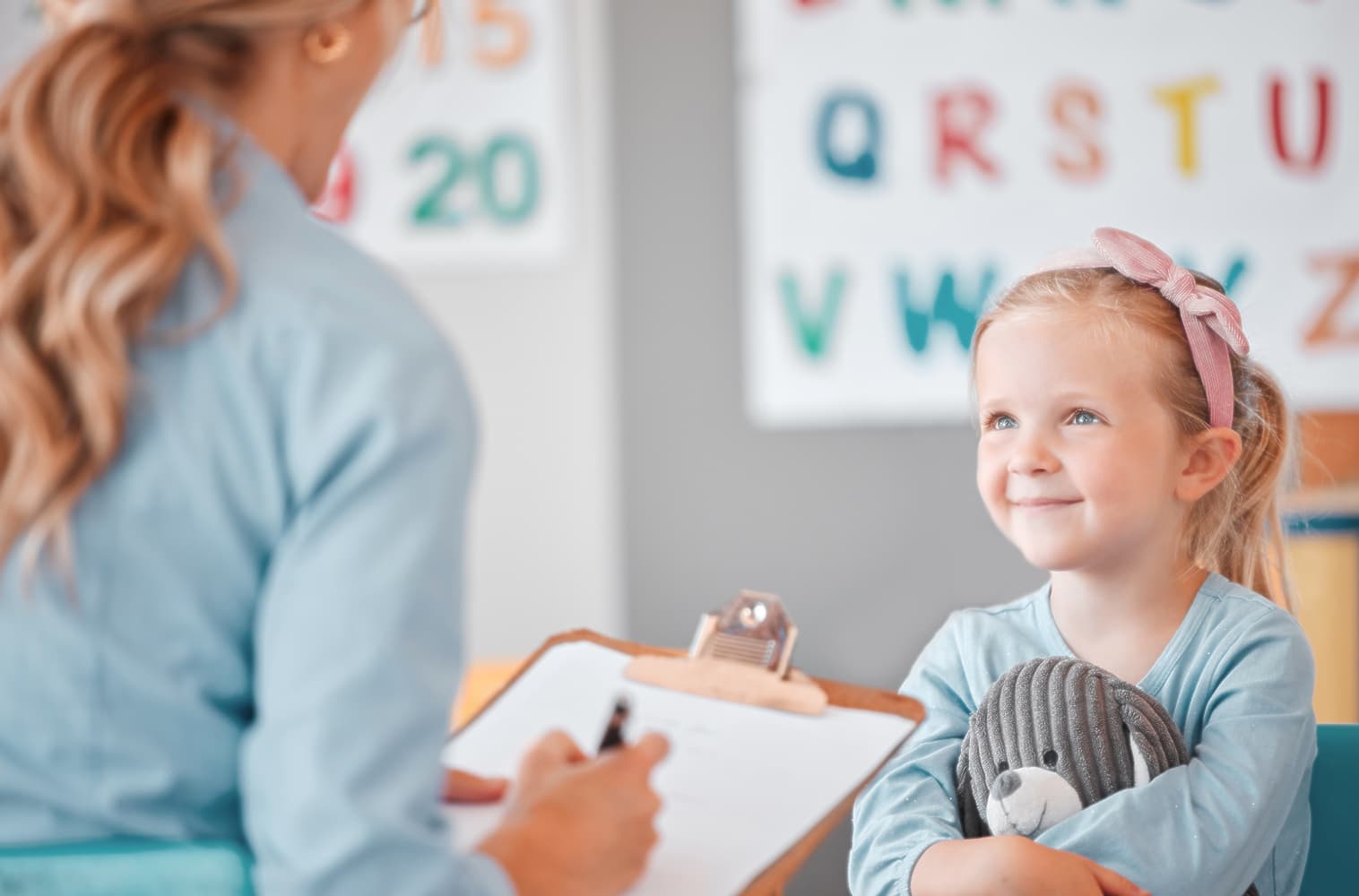 Young girl smiling and hugging a stuffed animal while looking at a speech therapist holding a clipboard, in a colorful speech and language therapy room.
