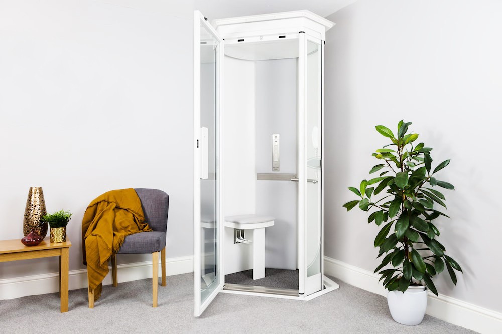 Elevate AVA Rise home lift in white with door open installed in a residential room corner — fully glazed cabin with interior fold-down seat and control panel visible