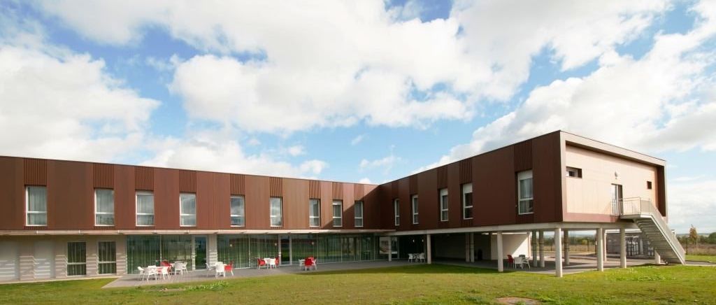 Modern healthcare facility exterior with brown corrugated cladding, elevated architectural feature on white pillars, and outdoor seating scattered across a manicured lawn.