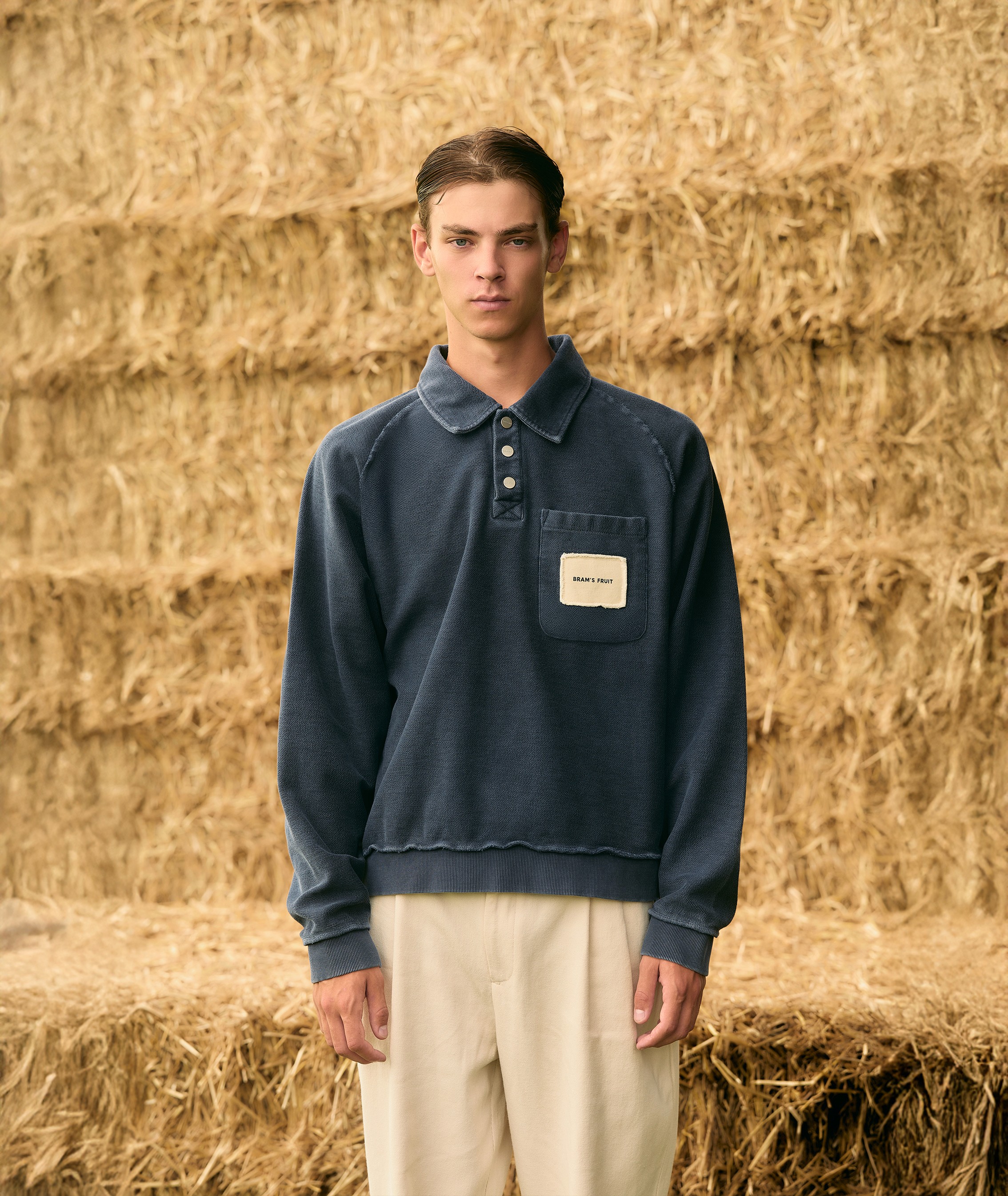 Plan américain devant des bottes de foin, le mannequin porte un polo délavé marine à manches longues de la marque Brams Fruit