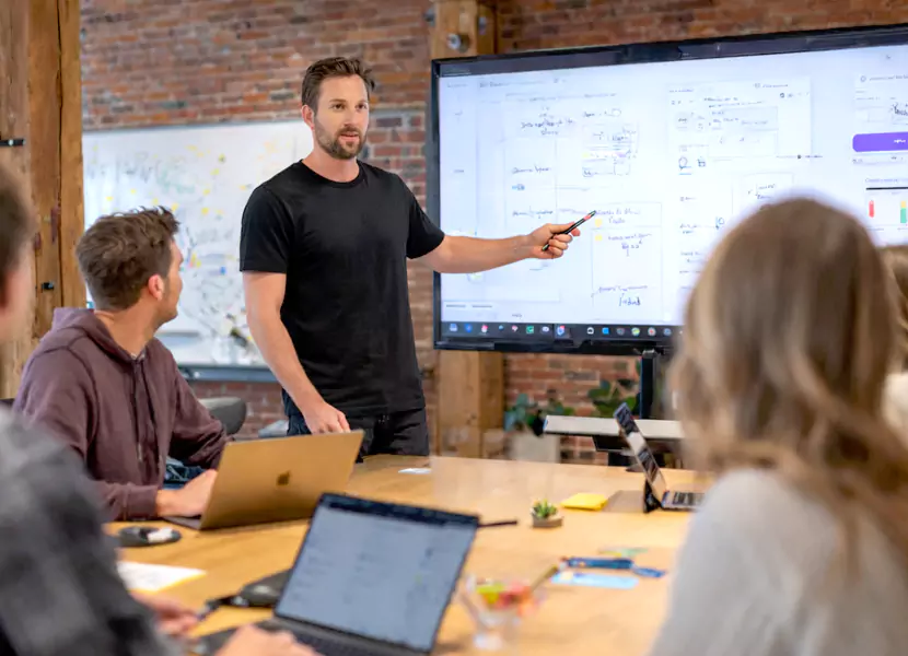 A man presents data on a screen to a group of colleagues in a modern meeting room. Laptops are on the table.