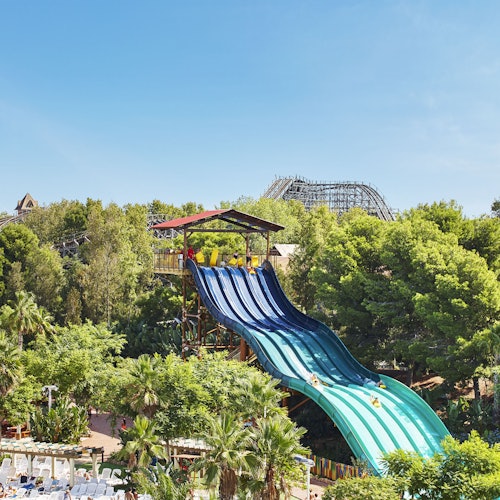 Multiple water slides with people descending from a tall platform, surrounded by lush greenery and a visible wooden roller coaster in the background.