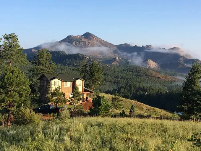 Luxury mountain estate with log cabin architecture, green meadow foreground, dense pine forest and misty Rocky Mountain peaks at sunrise - scenic outdoor wedding venues in Colorado