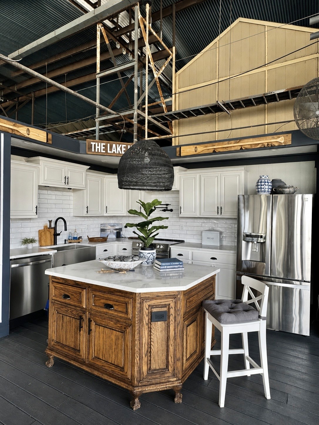 A modern farmhouse kitchen featuring white cabinets, a marble-topped wooden island, stainless steel appliances, and industrial-style accents such as overhead metal beams and a large woven pendant light.